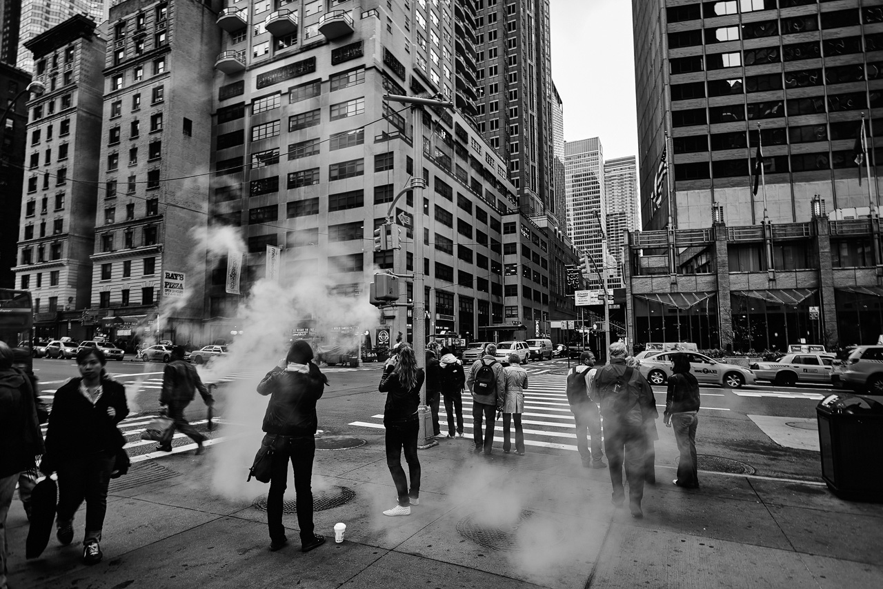 Menschen stehen auf einer Stra&szlig;e in Manhattan, Rauch steigt aus einem Kanaldeckel auf in New York von Fotograf Dominique Hammer